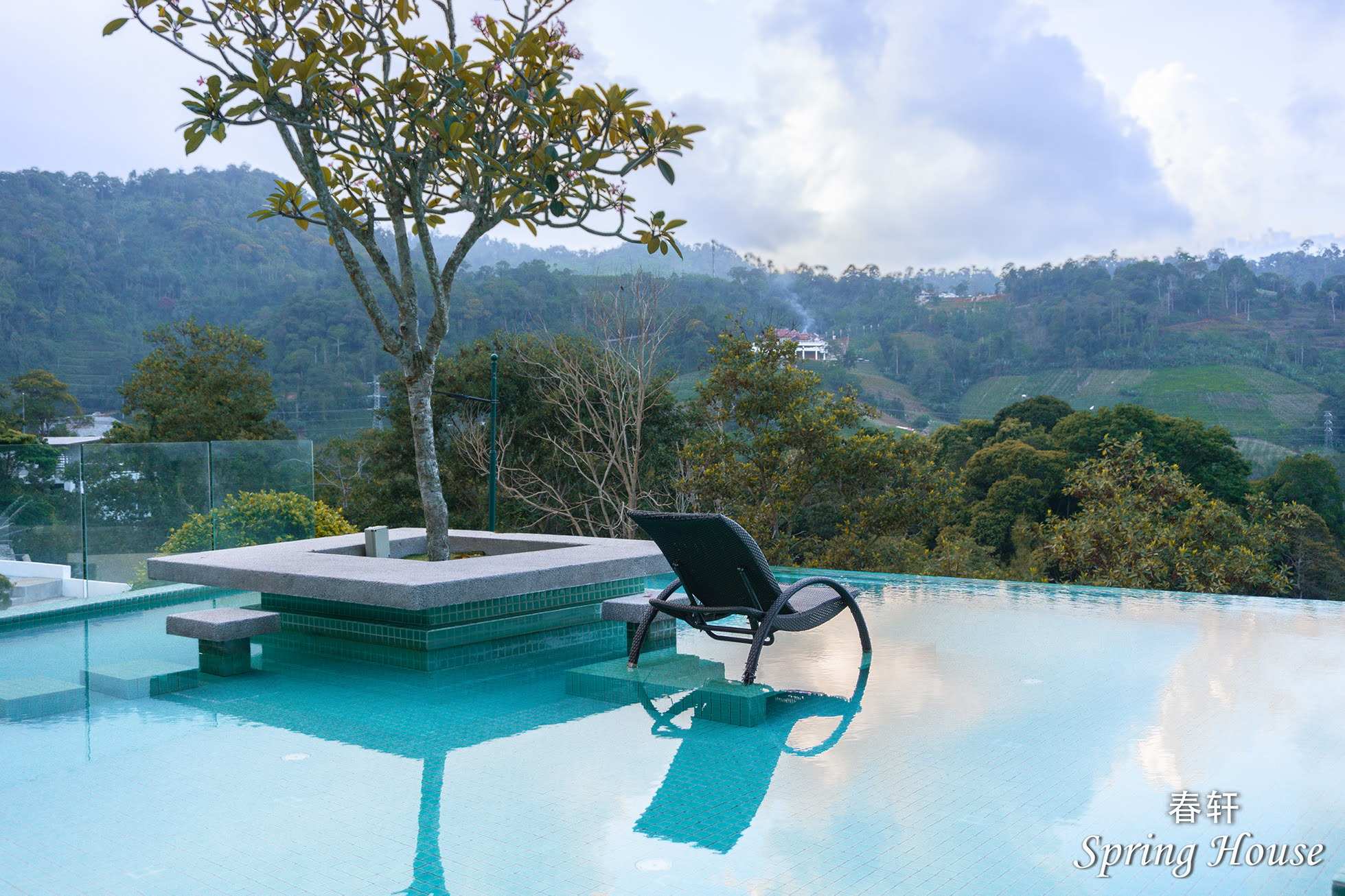 Infinity pool corner with a wicker lounge chair standing in the water, a frangipani planter island and the misty valley beyond.