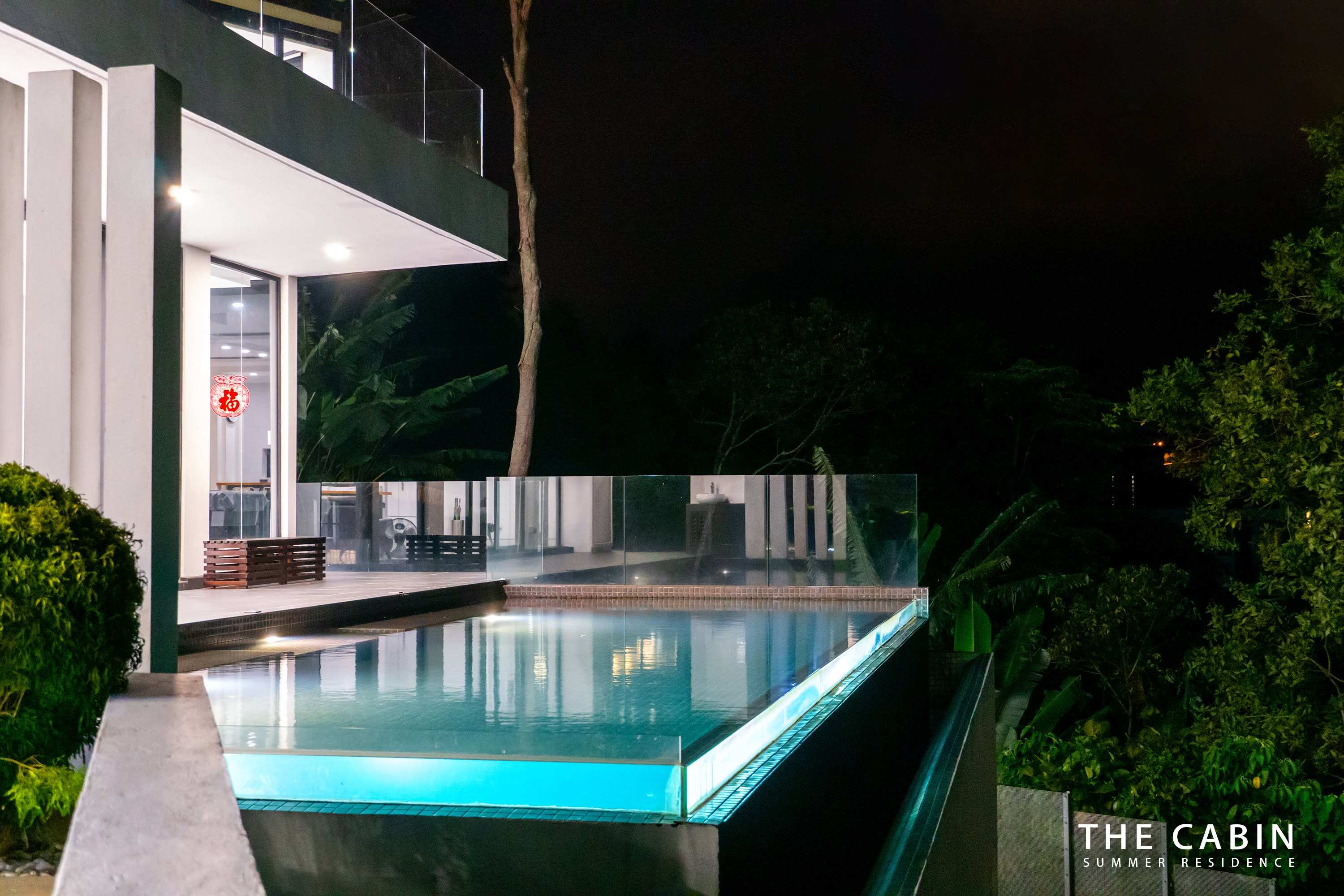 The villa at night above the infinity pool, warm interior light behind the glass wall.