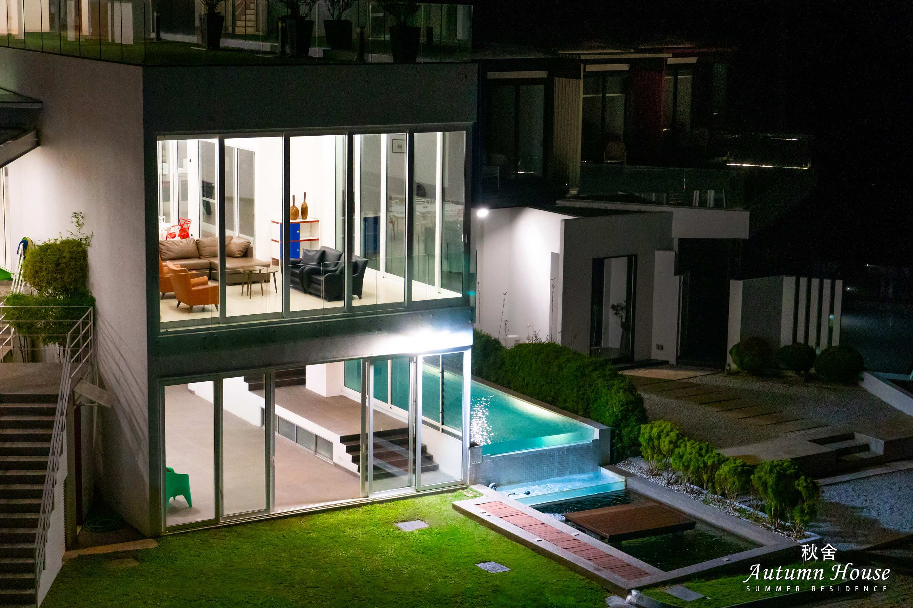 The villa at night: a two-storey glass facade lit from within, opening onto the illuminated infinity pool.
