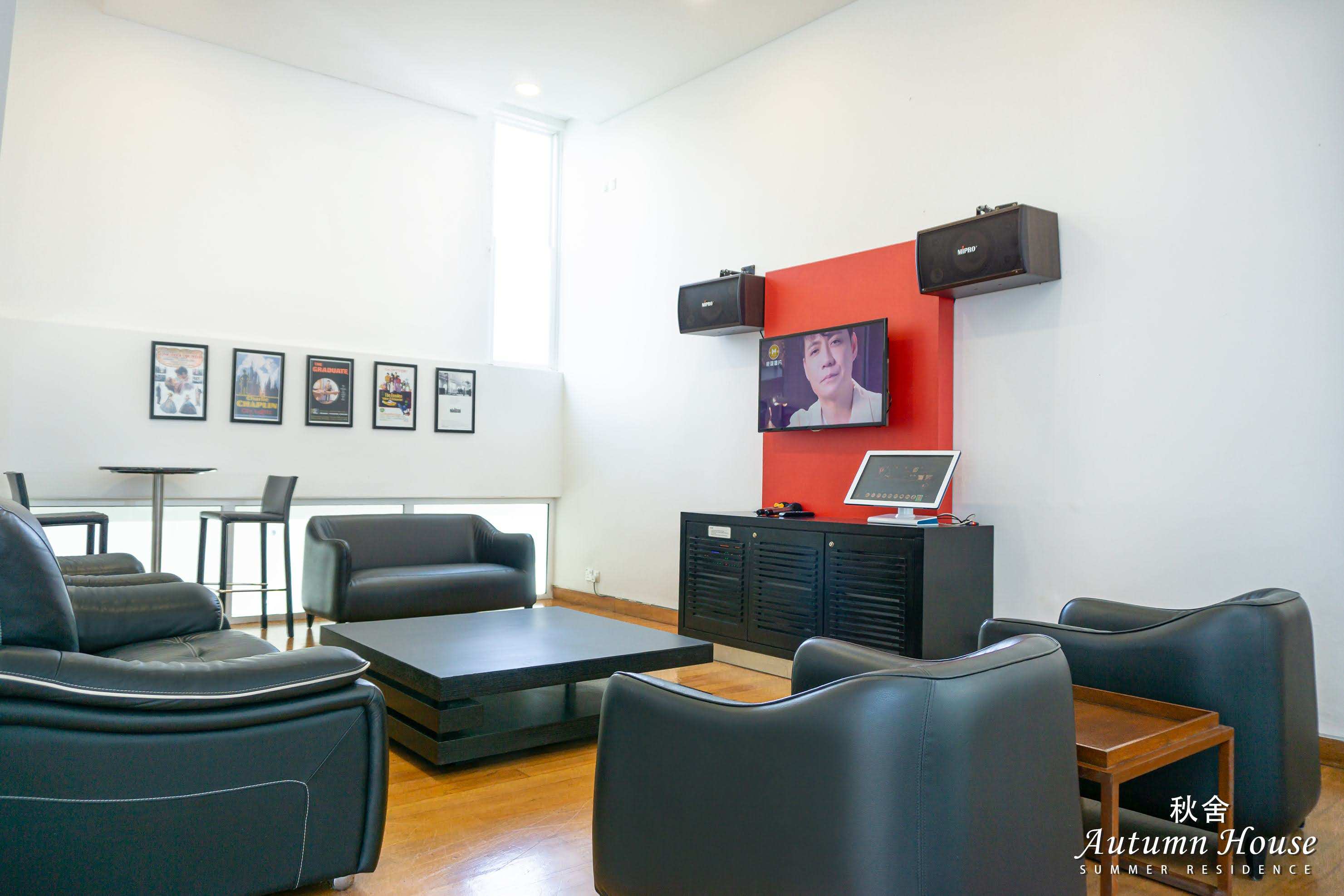 Karaoke room from floor level: black leather sofas facing the red wall with screen and speakers, framed posters alongside.