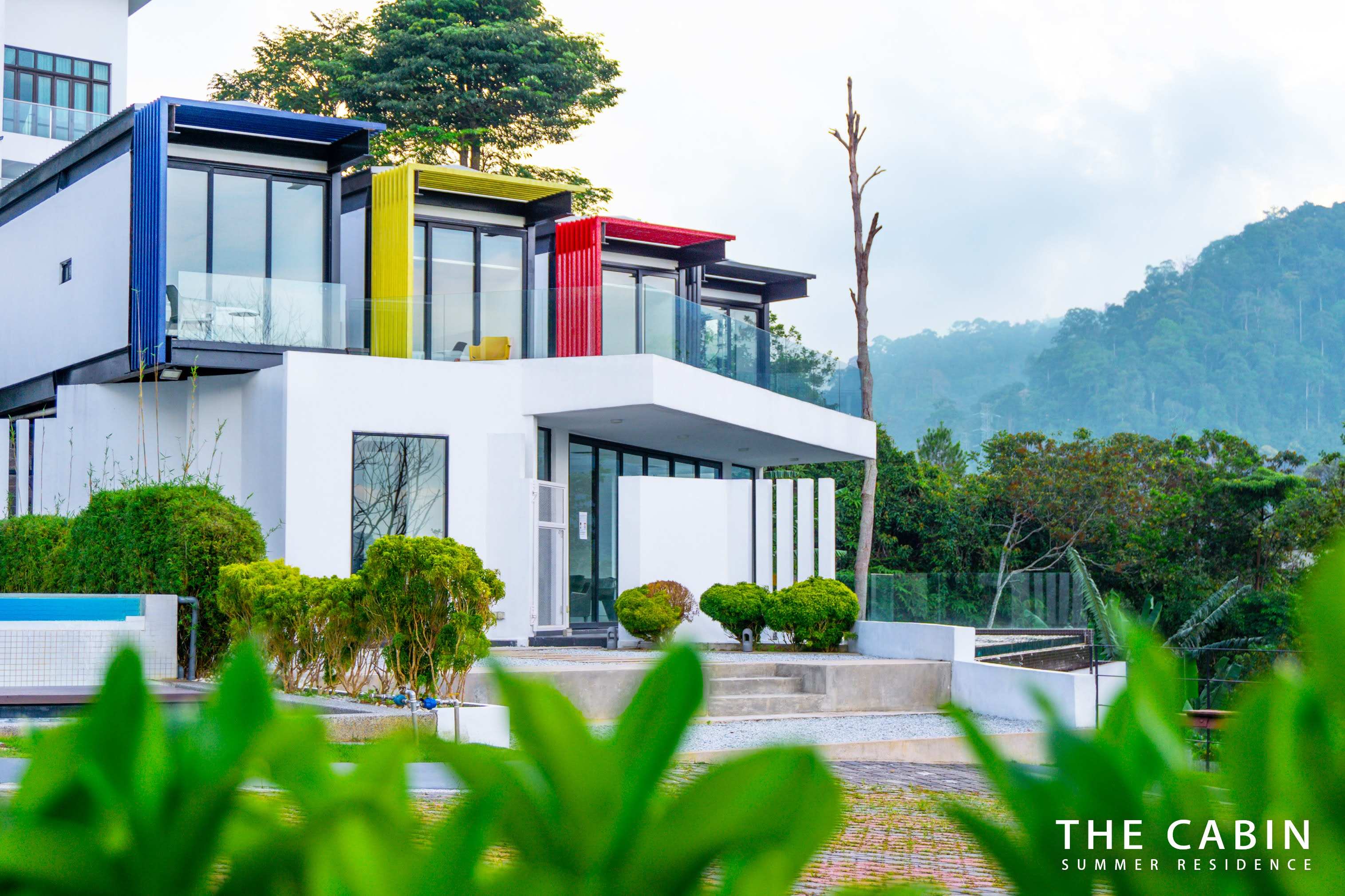 Front elevation of the villa with its blue, yellow and red slat balcony accents against the mountain backdrop.