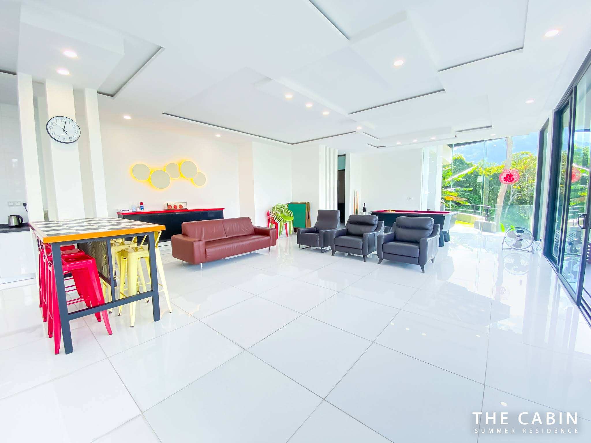 The open-plan common hall: red sofa and leather armchairs in front, pool table and karaoke doorway behind, glass doors to the pool deck.
