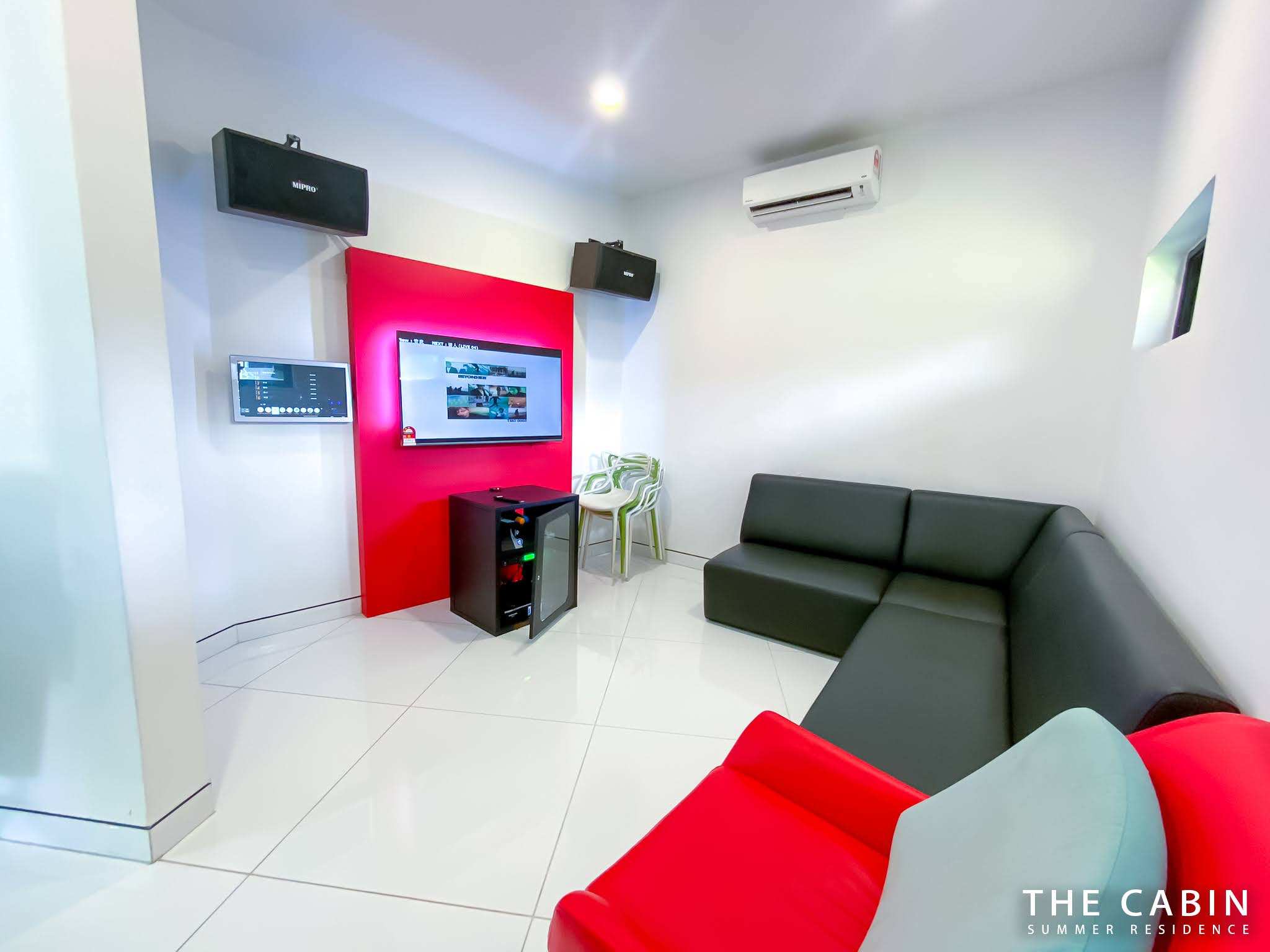 Karaoke room: red accent wall with the screen and song selector, wall-mounted speakers and a black leather corner sofa.