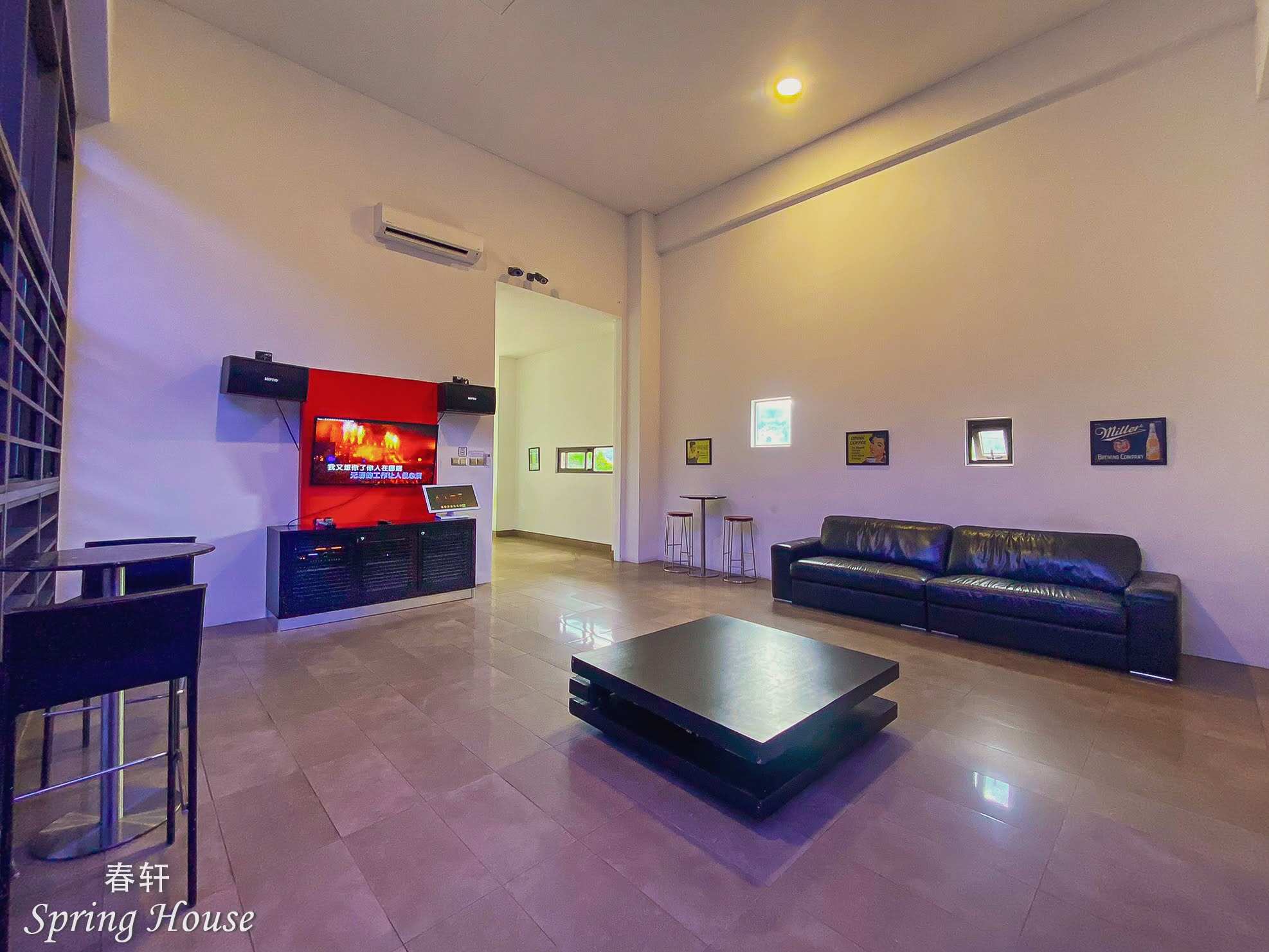 Karaoke lounge: red feature wall with the screen, black leather sofa, bar stools and retro sign art under a high ceiling.