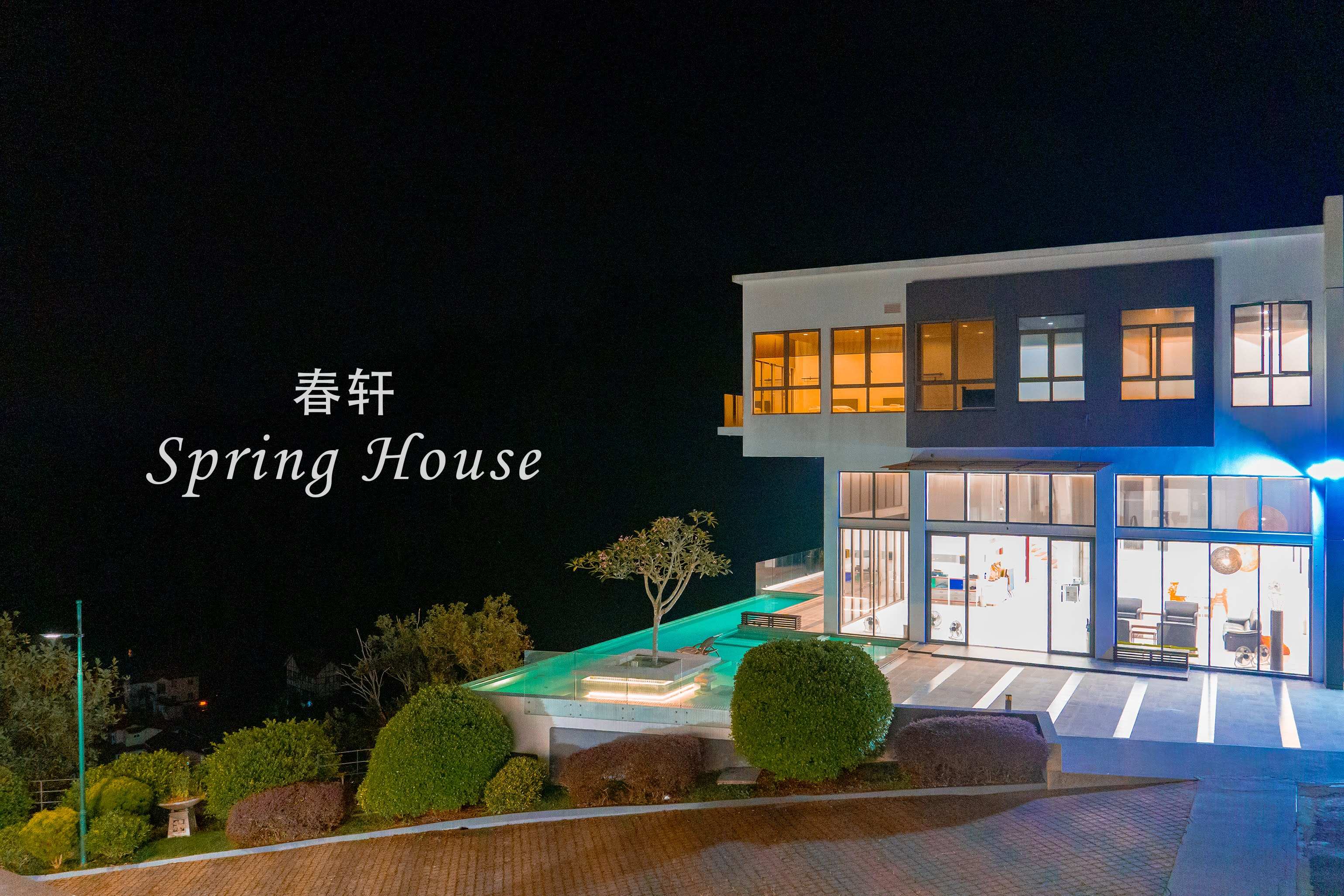 Night exterior with the glowing turquoise pool in front and the lit lounge visible through the glass walls.