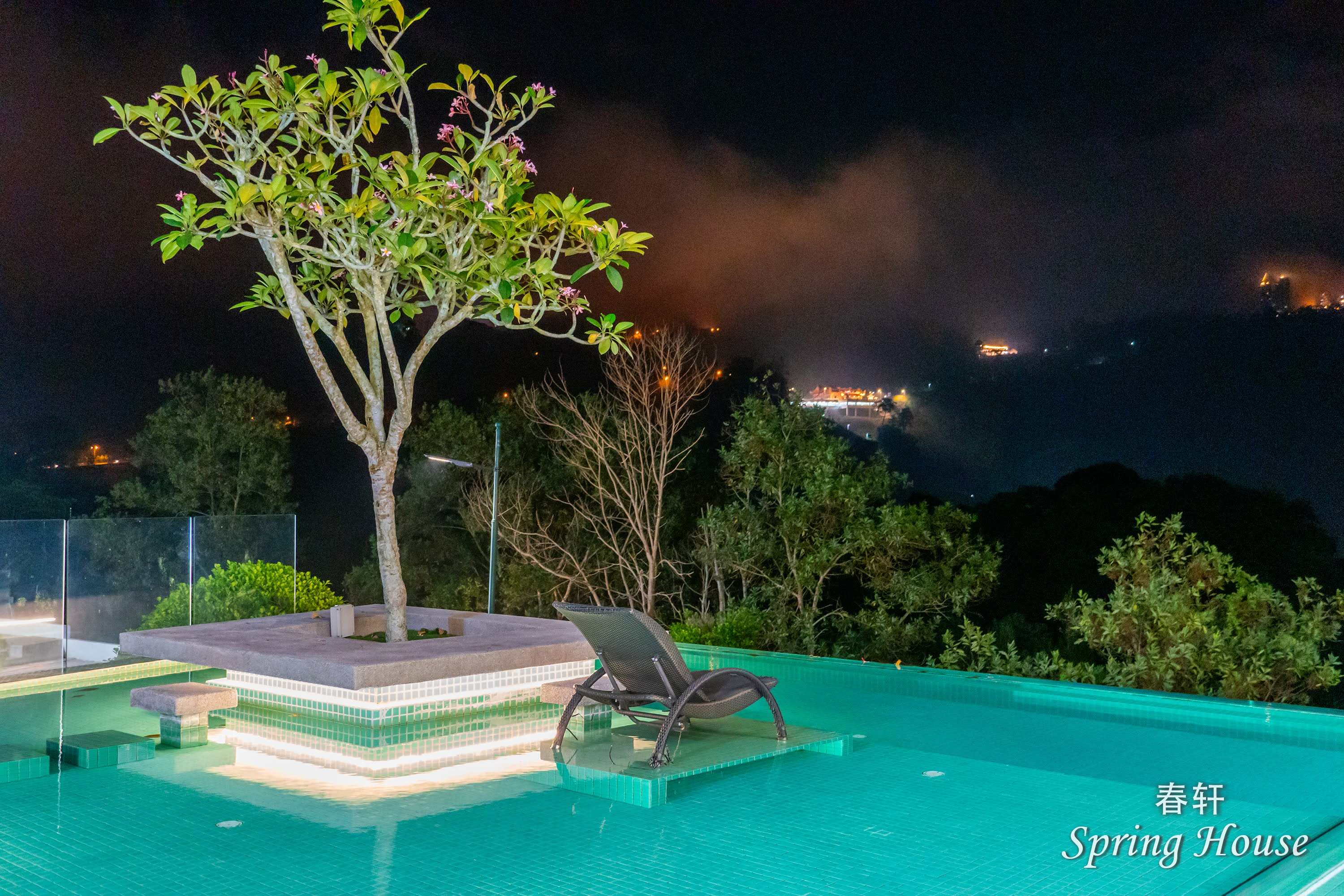 Pool-level night shot of the tree island and lounge chair, distant hillside lights behind.