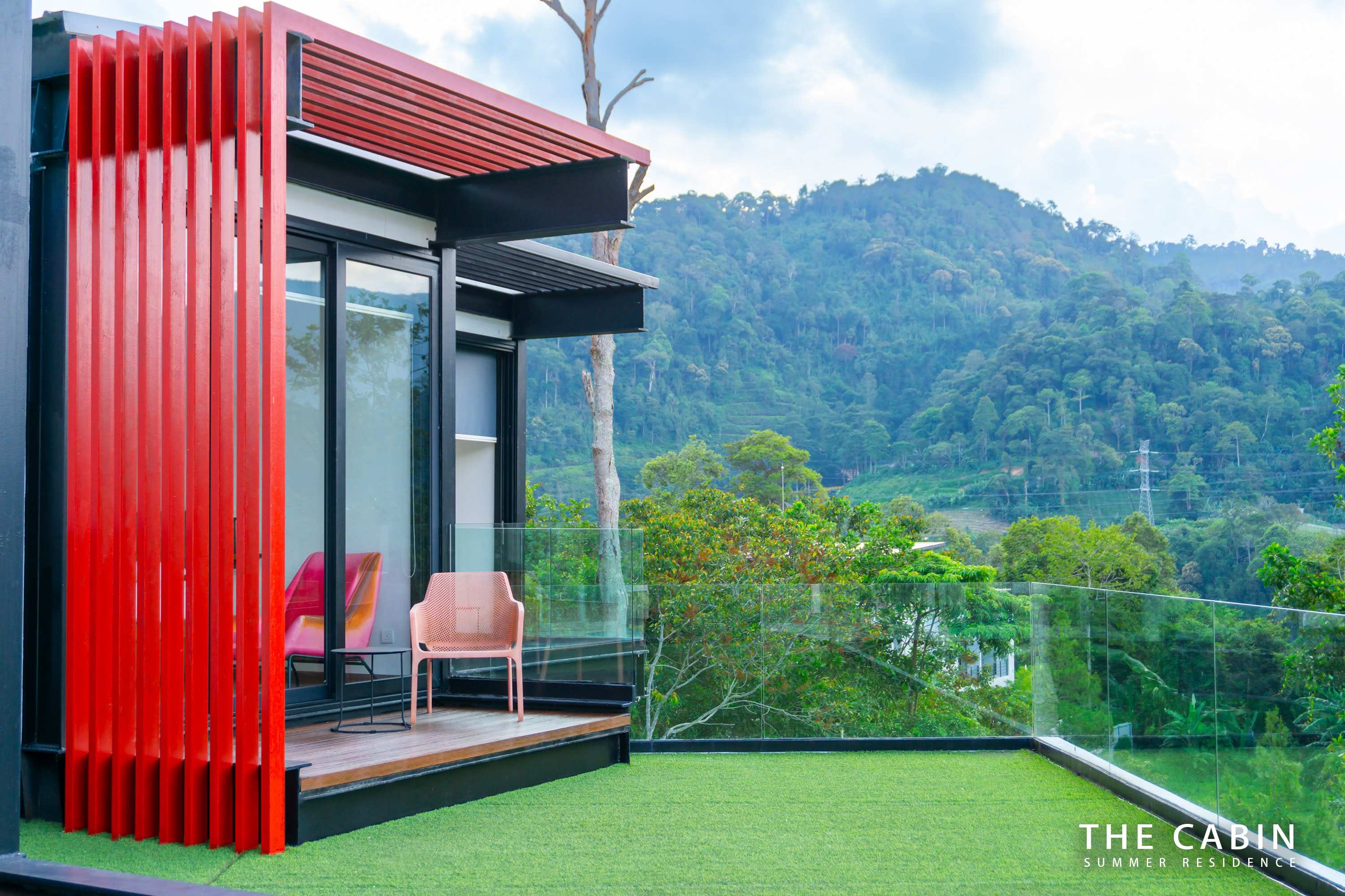 A cabin module clad in red slats, with coral balcony chairs and artificial turf over the mountain view.